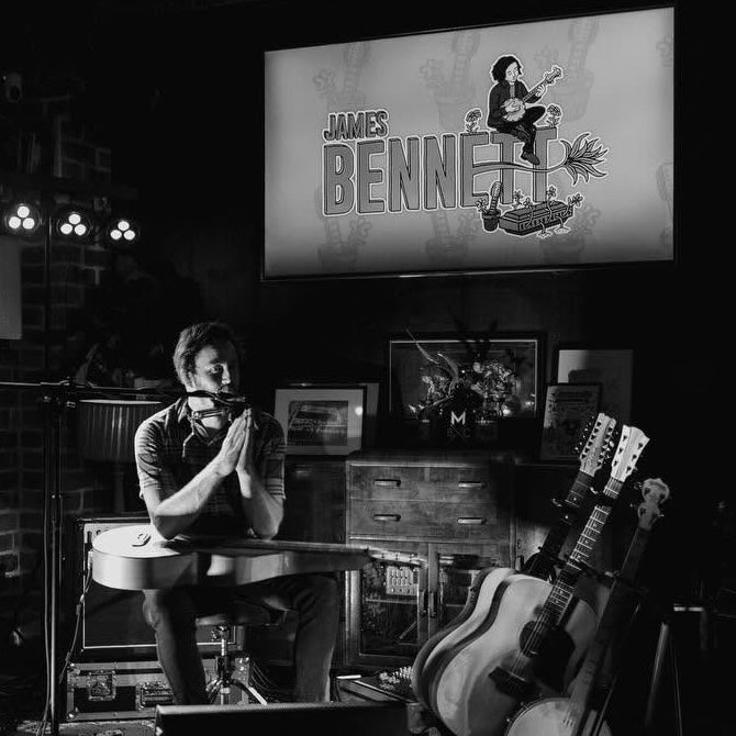 A black and white photograph of a live music performance in a dimly lit venue. JAMES BENNETT is visible at a table, with acoustic guitars leaning against a stand, a brick wall on the left, and a large screen on the wall displaying the text 'JAMES BENNETT'.